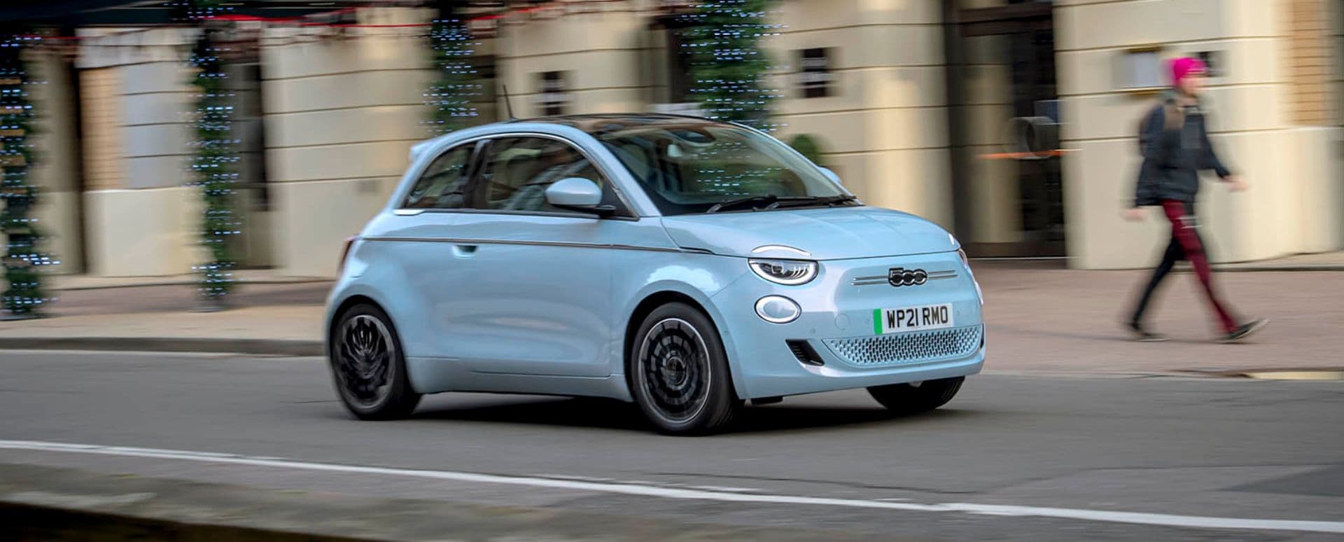 Baby blue Fiat 500e driving in the city with a woman wearing a pink beanie walking by