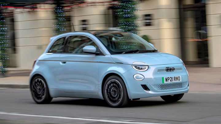 Baby blue Fiat 500e driving in the city with a woman wearing a pink beanie walking by