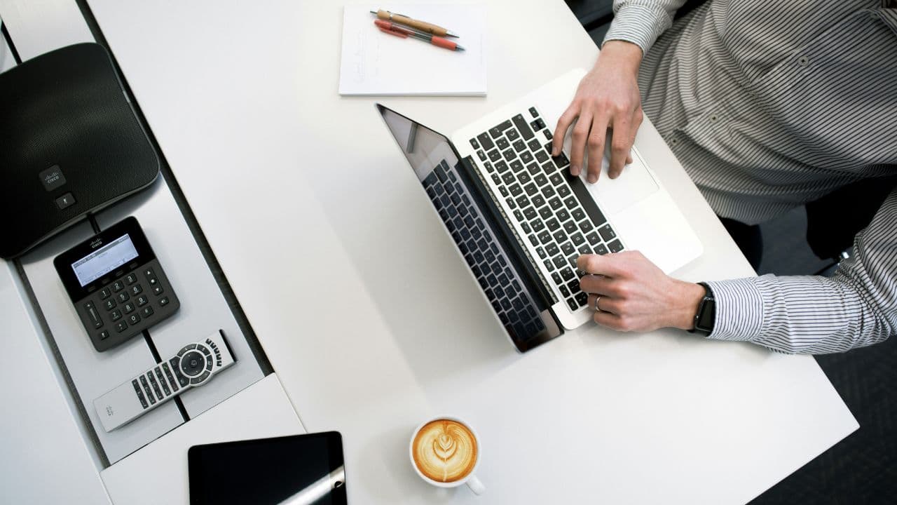 Man sitting at a desk typing on his laptop with a mug of coffee sitting in front of the laptop