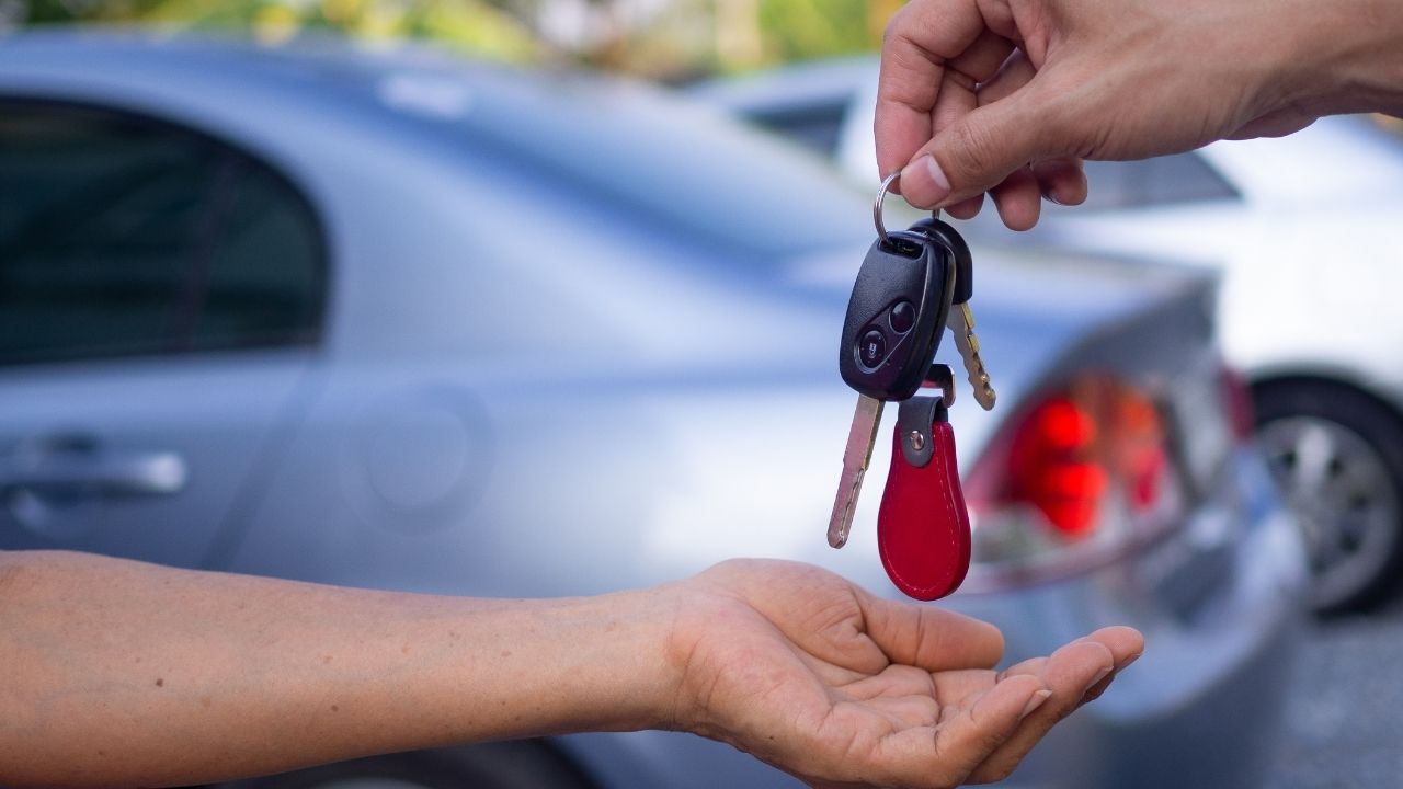 A person handing a car key to another person in front of a silver car