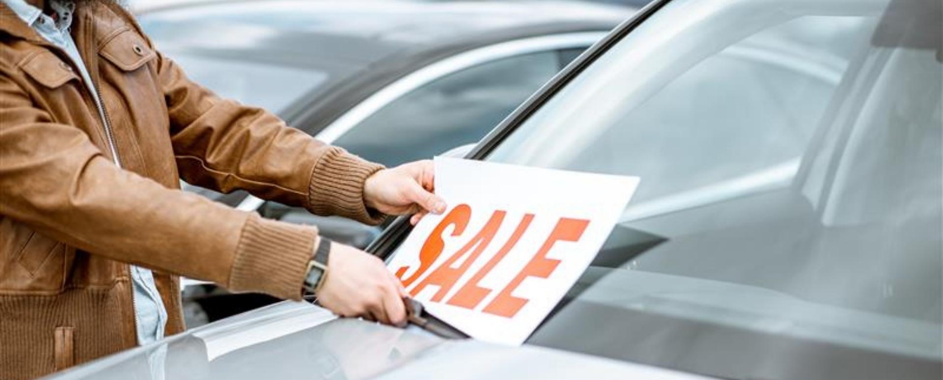 A person wearing a brown jacket puts a sale sign on a vehicle's windshield