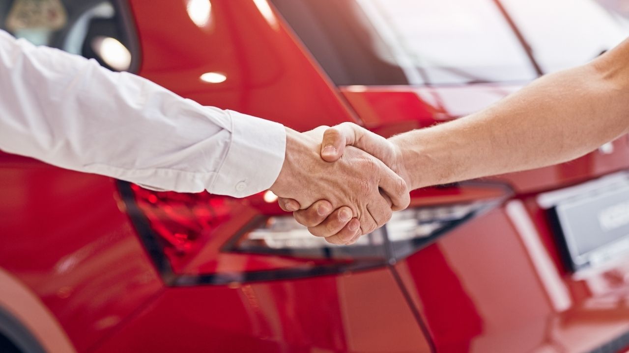 Two people shacking hands in front of a red car