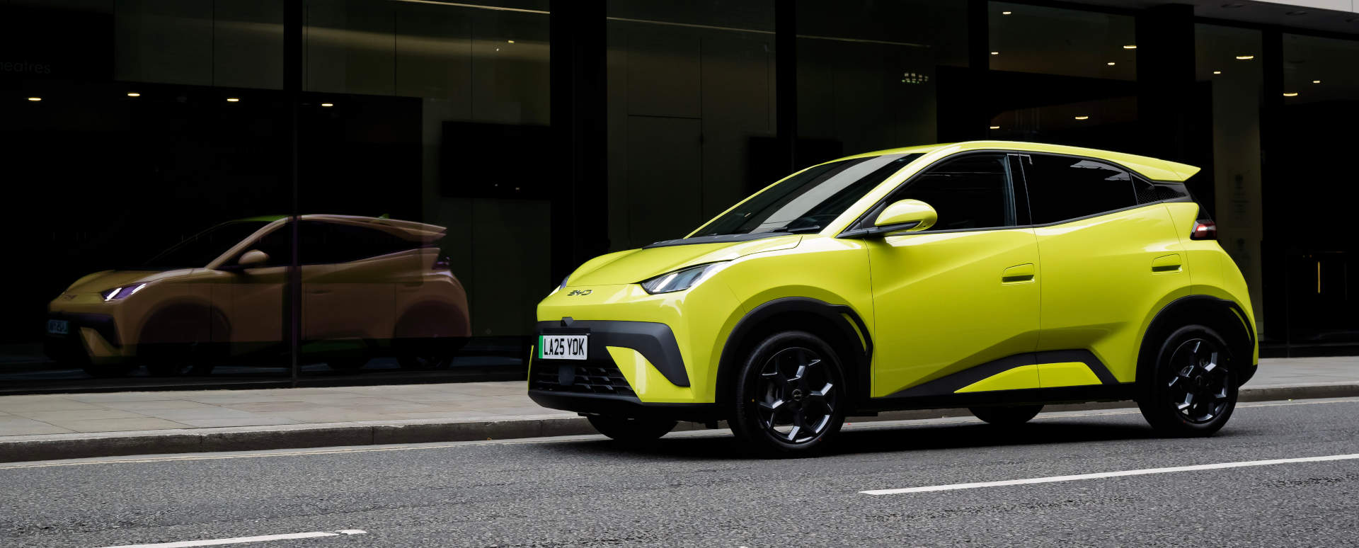 Yellow BYD Dolphin Surf Driving Reflected in Window of Building