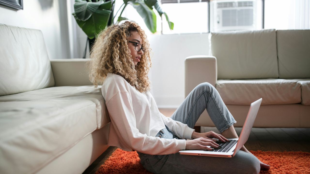 Woman in white shirt and jeans leaning on a couch whilst sitting on the floor of a living room and typing on her laptop