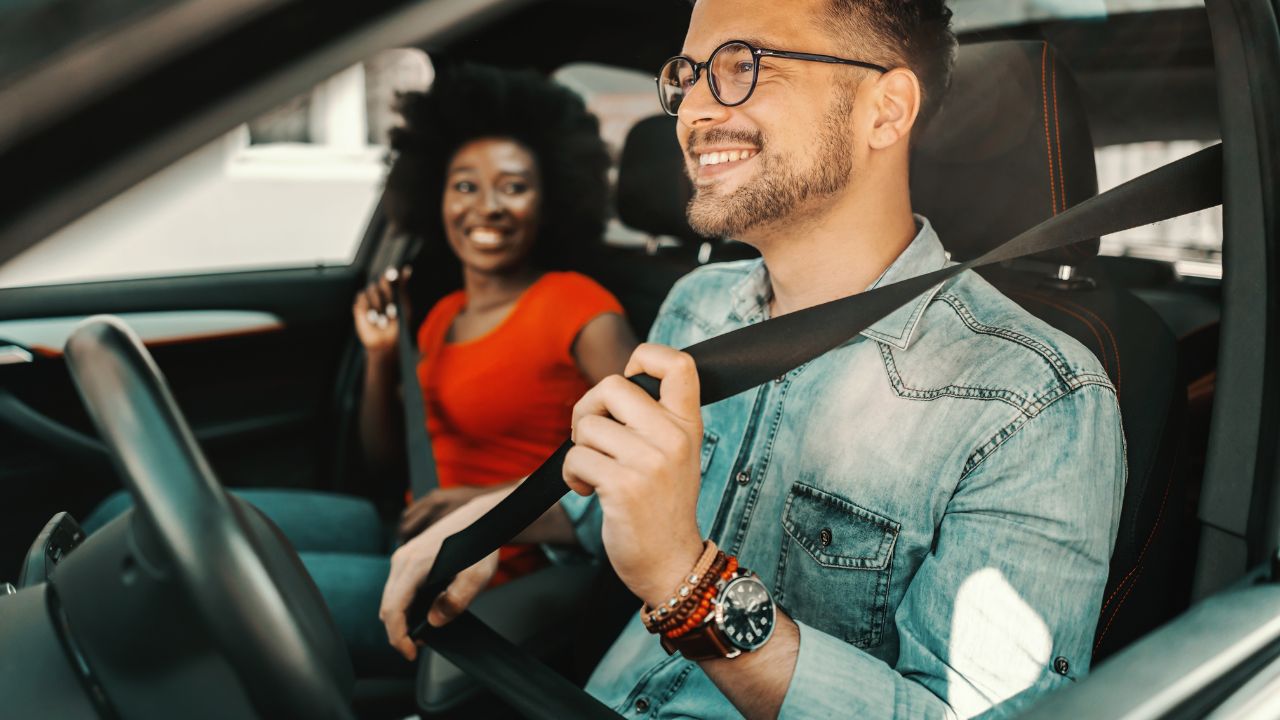 Woman wearing an orange shirt seating in the passenger seat of a car putting on her seatbelt. Man wearing a jeans shirt seating in the driver's seat putting on seatbelt.