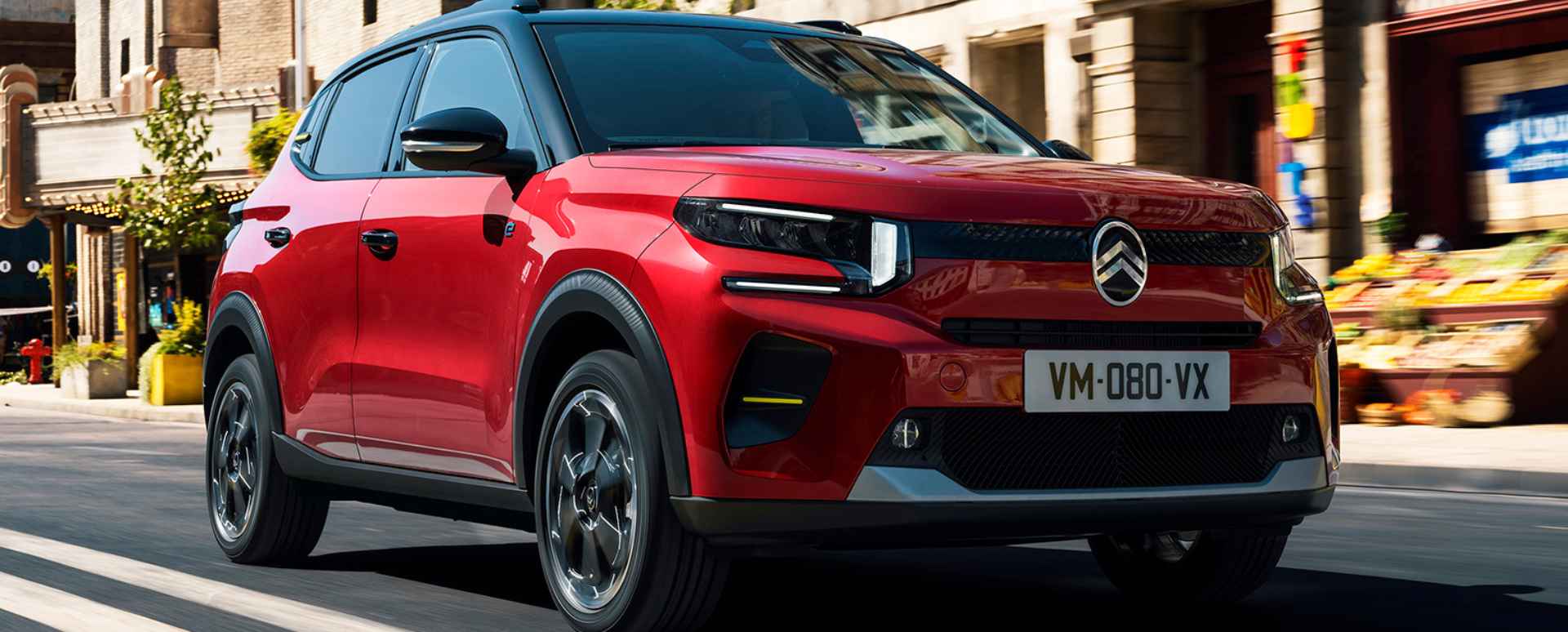 Red Citroen C3 driving on the street with shops on the side
