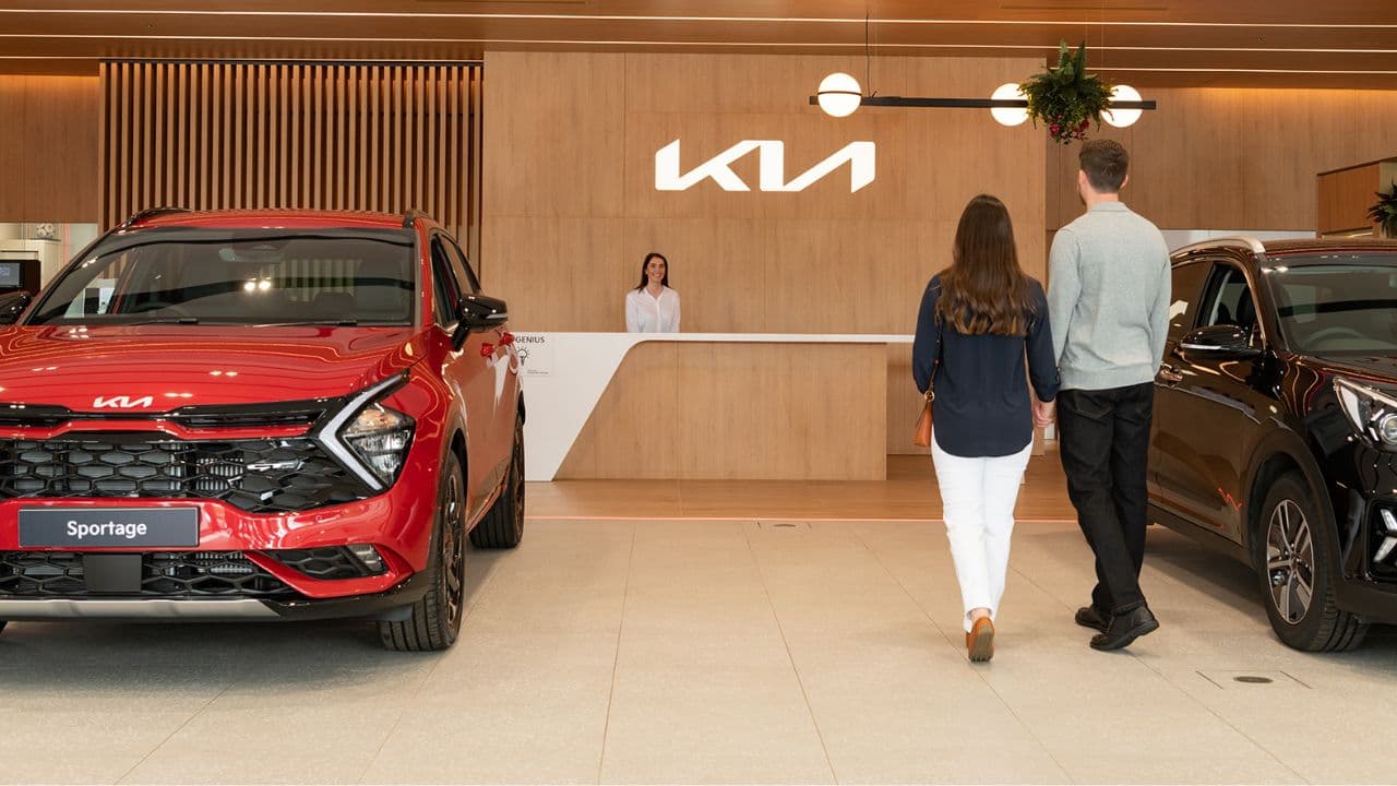 Kia dealership with a red Kia car parked on the left and a black car parked on the right. A couple is walking up the counter.