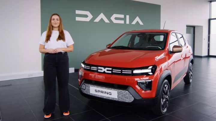 Red Dacia Spring parked in a Dacia showroom with a woman wearing a pair of black trousers and white t-shirt standing next to it