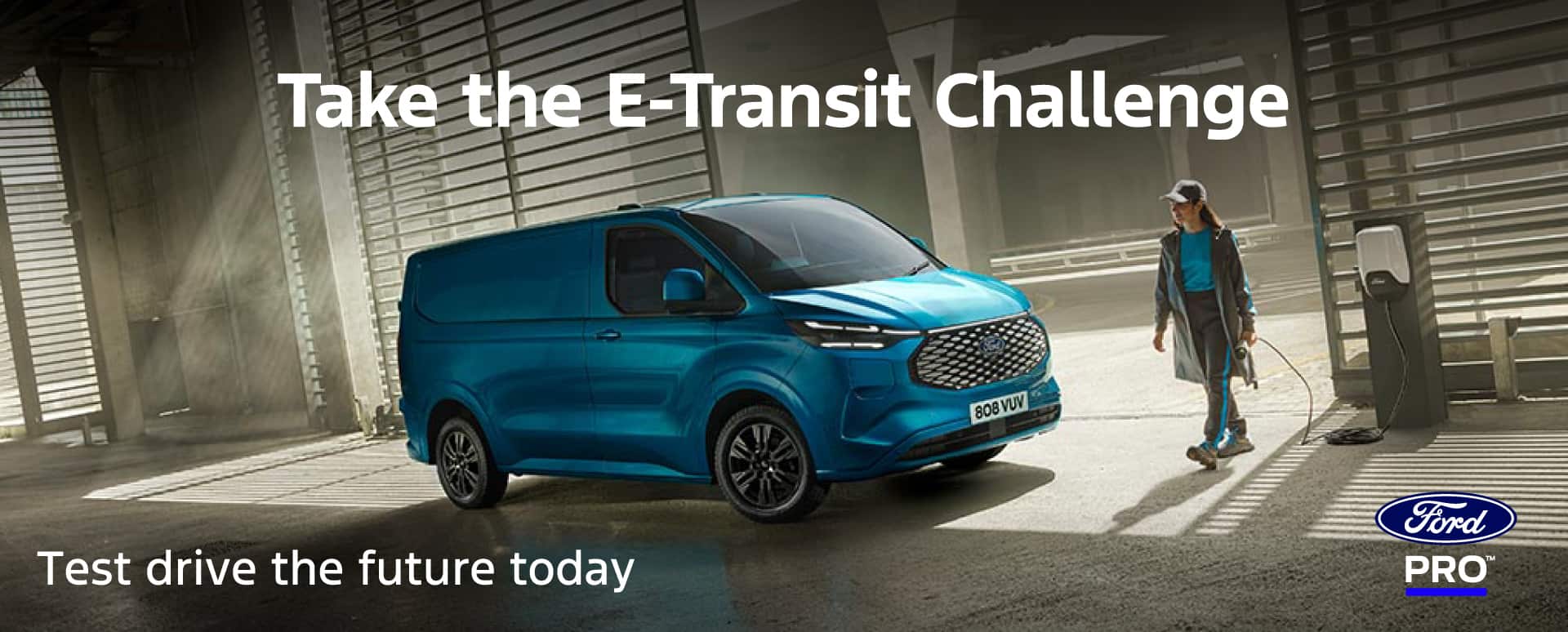 A blue Ford E-Transit parked in front of roll-up doors and a woman wearing the same shade of blue as the van is holding a charger and walking towards the socket