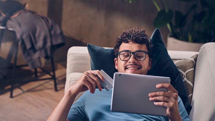 Man lying on the couch with the head on the cushion while holding a credit card and a tablet