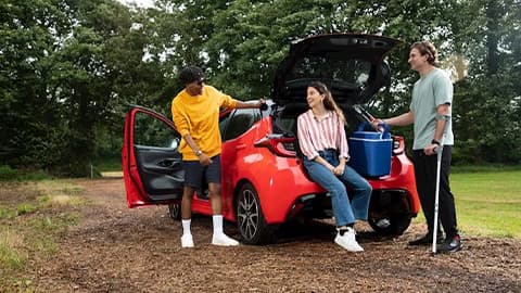 A group of three young adults gathered around a red Toyota Yaris in a country park