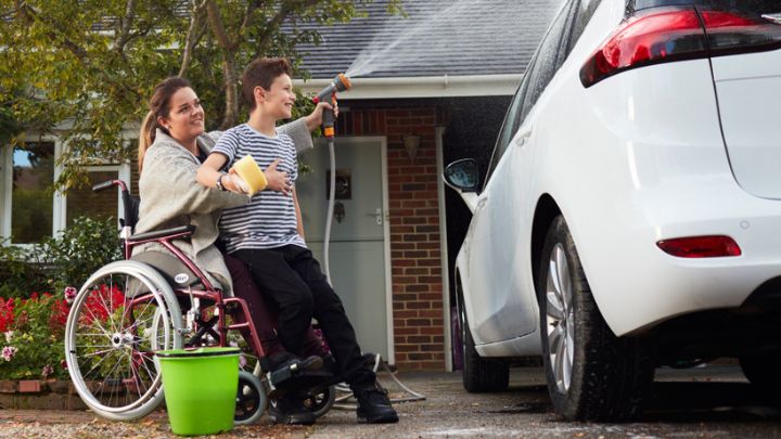 Woman on a wheelchair helping her son wash a white car parked in her drive way with a hose