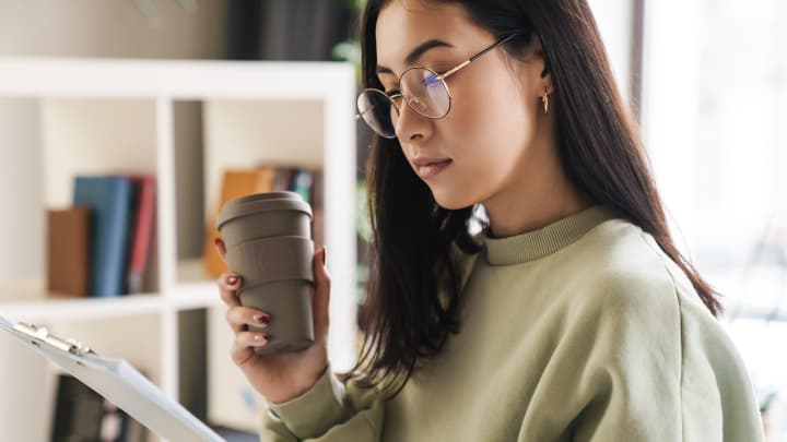 Dark haired woman looking at documents whilst holding a reusable coffee cup