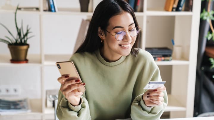 Dark haired woman holding her smartphone on one hand and reading her credit card details whilst holding it on her other hand