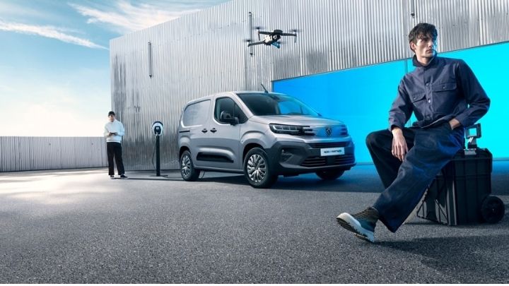 Side view of a silver Peugeot e-Partner parked in front of a grey wall with one man sitting on a suitcase on the left handside