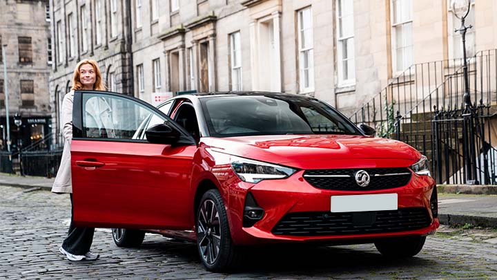 Red Vauxhall Corsa parked in city, with lady standing next to it with the driver's door open